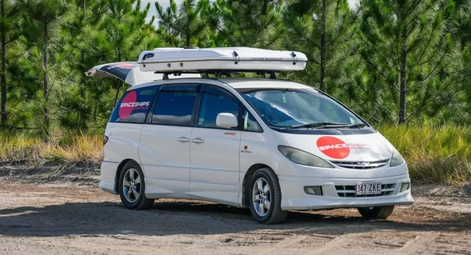 weißer Minicamper mit Dachbox auf Sand vor Bäumen