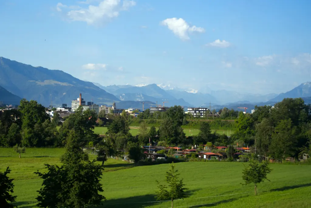 Malerische Landschaft mit grünen Feldern und Bäumen im Vordergrund; im Hintergrund eine von Bergen umgebene Stadt unter blauem Himmel mit vereinzelten Wolken.
